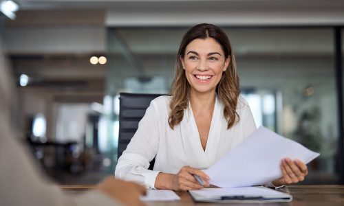 Smiling mature business woman hr holding cv document at job interview. Happy mid aged professional banking manager or lawyer consulting client sitting at workplace in corporate office meeting.