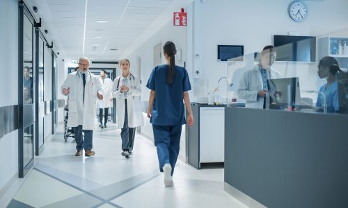 Diverse Male and Female Medical Professionals Navigate a Busy Hospital Corridor and Registration Area, Having Conversations About Healthcare. Male Nurse Assists a Patient in a Wheelchair